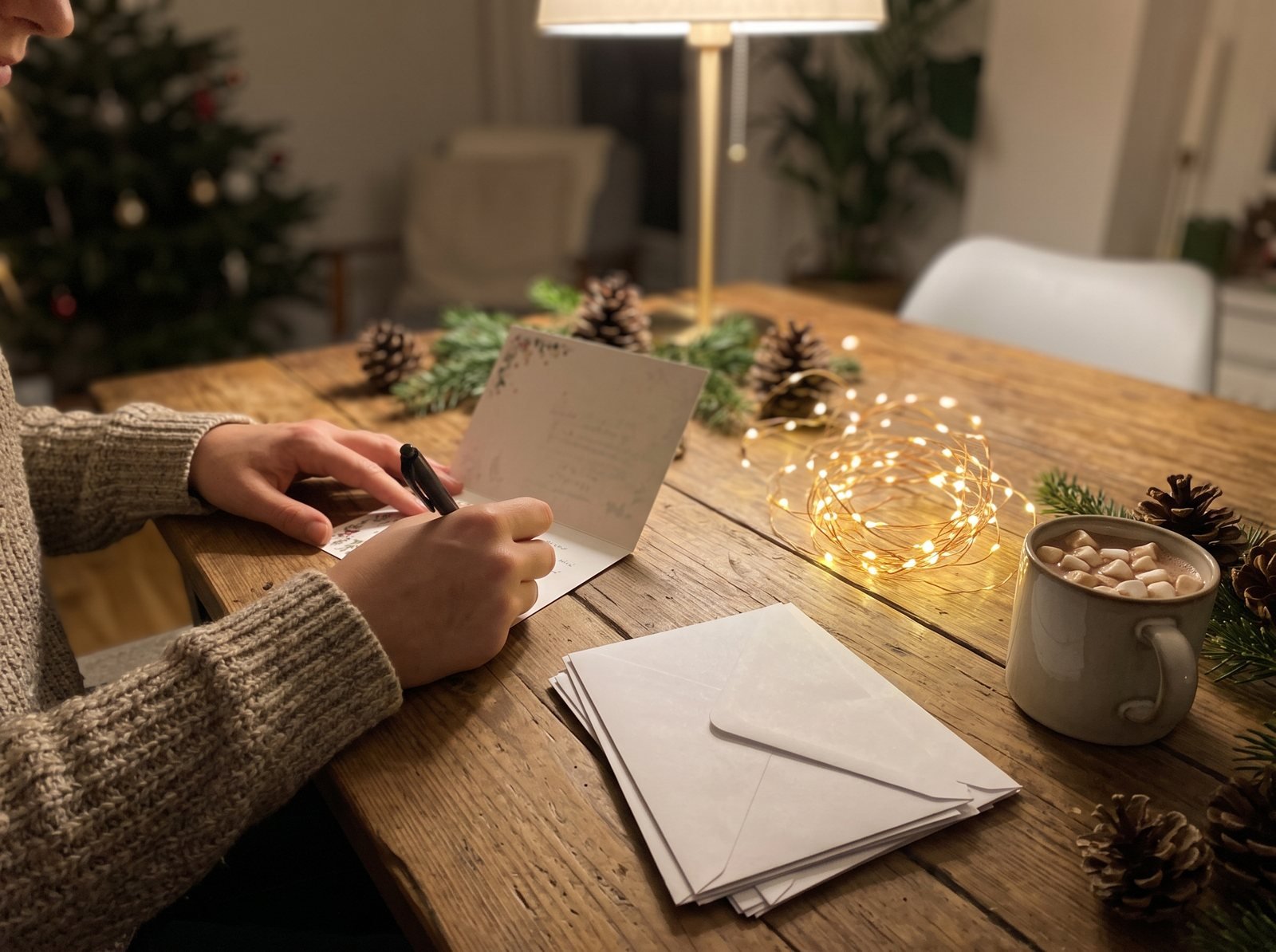 Hands writing a holiday card at a table with envelopes, string lights and cocoa